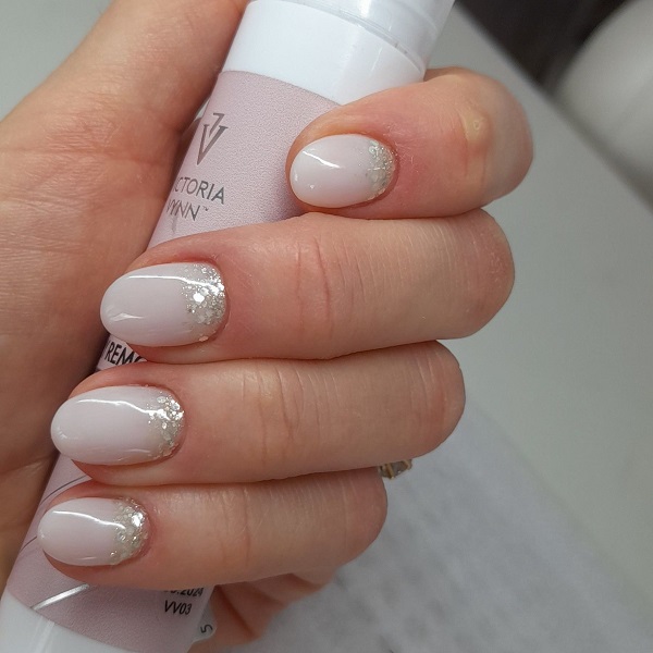 Paznokcie hybrydowe w Rzeszowie  Na zdjęciu widzimy rękę z pięknie wykonanym manicure hybrydowym. Paznokcie są pomalowane na elegancki, jasnoróżowy kolor z subtelnym zdobieniem w postaci brokatowych akcentów przy skórkach. Taki manicure hybrydowy jest trwały i estetyczny, idealny na różne okazje. Dłoń trzyma produkt do pielęgnacji paznokci marki Victoria Vynn, co sugeruje profesjonalne podejście do zabiegu i użycie wysokiej jakości kosmetyków. Tego typu usługa jest dostępna w salonach kosmetycznych w Rzeszowie, oferując klientkom piękne i zadbane dłonie na dłużej