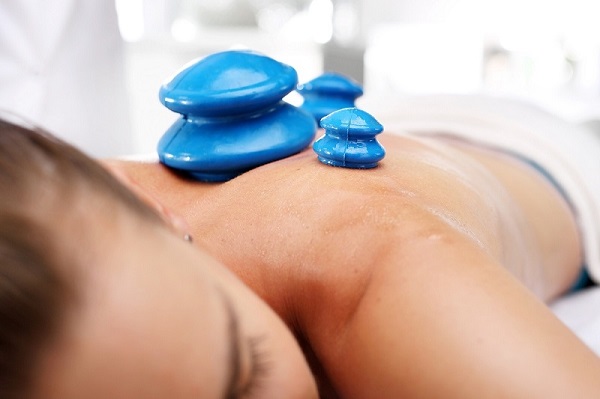 Masaż bańką chińską - Na zdjęciu widoczna jest osoba leżąca na brzuchu, poddawana masażowi bańką chińską. Na plecach pacjenta umieszczone są trzy niebieskie bańki, które tworzą próżnię i pomagają w rozluźnieniu mięśni oraz poprawie krążenia. Tego typu masaż jest stosowany do redukcji bólu, eliminacji toksyn z organizmu oraz poprawy ogólnego samopoczucia. Pacjentka jest owinięta ręcznikiem, co sugeruje, że zabieg odbywa się w profesjonalnym gabinecie.