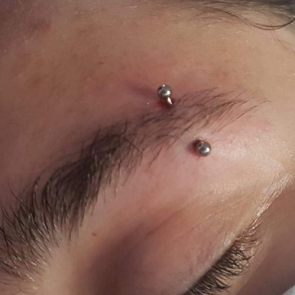 Przekłuwanie brwi - Zdjęcie przedstawia zbliżenie na brew osoby z kolczykiem umieszczonym w jej górnej części. Kolczyk składa się z dwóch srebrnych kulek, które są widoczne po obu stronach przekłucia. Skóra wokół miejsca przekłucia jest lekko zaczerwieniona. Brew jest naturalna i gęsta, a kolczyk dodaje jej charakteru.