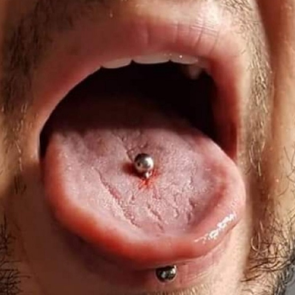 Przekłuwanie języka - Zdjęcie przedstawia zbliżenie na usta osoby z wyciągniętym językiem, na którym znajduje się kolczyk. Kolczyk jest umieszczony w centralnej części języka i ma srebrną kulkę na obu końcach. Widać delikatne zaczerwienienie wokół miejsca przekłucia. Dodatkowo, na dolnej wardze widoczny jest drugi kolczyk z ciemniejszą kulką.