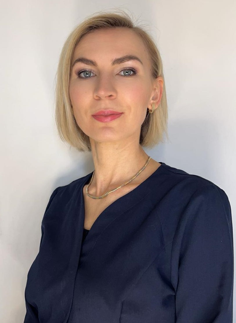 Kobieta z ciemnymi włosami ubrana w biały uniform, stojąca na tle jasnej ściany. Ma na sobie naszyjnik i delikatny makijaż. Uśmiecha się delikatnie, wyglądając profesjonalnie i przyjaźnie.