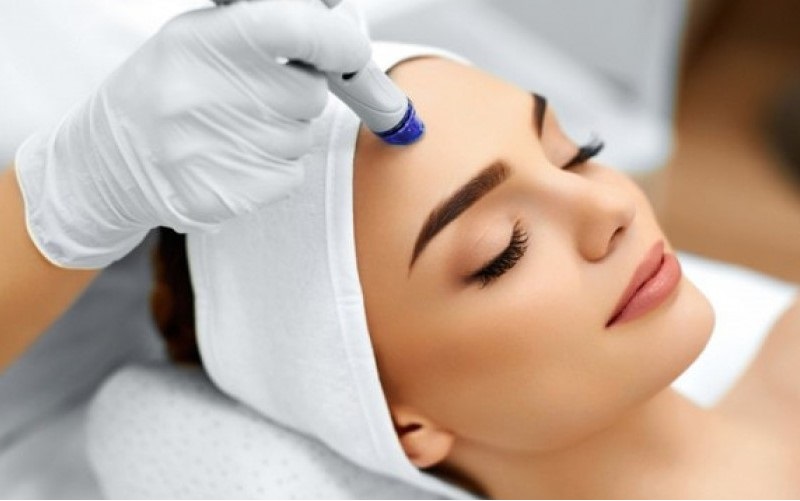 Na zdjęciu widać kobietę leżącą na plecach, zrelaksowaną, podczas profesjonalnego zabiegu kosmetycznego na twarzy. Specjalista w białych rękawiczkach aplikuje urządzenie na czoło pacjentki. Kobieta ma zamknięte oczy i wyraz spokoju na twarzy. Cała scena odbywa się w sterylnych, klinicznych warunkach, co sugeruje profesjonalną opiekę kosmetologiczną.